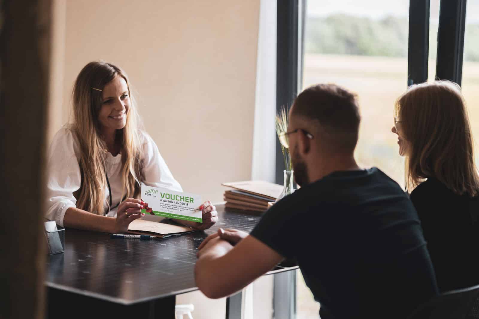Uśmiechnięta kobieta pokazująca voucher dwóm osobom podczas spotkania przy stole.