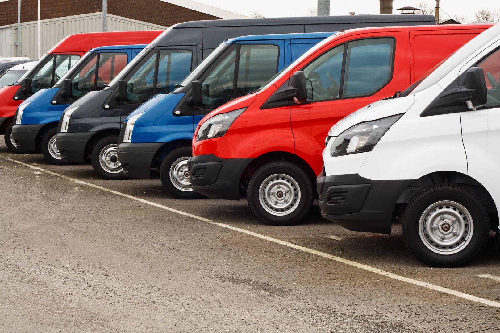 Rząd wielokolorowych samochodów dostawczych zaparkowanych po przekątnej na parkingu.