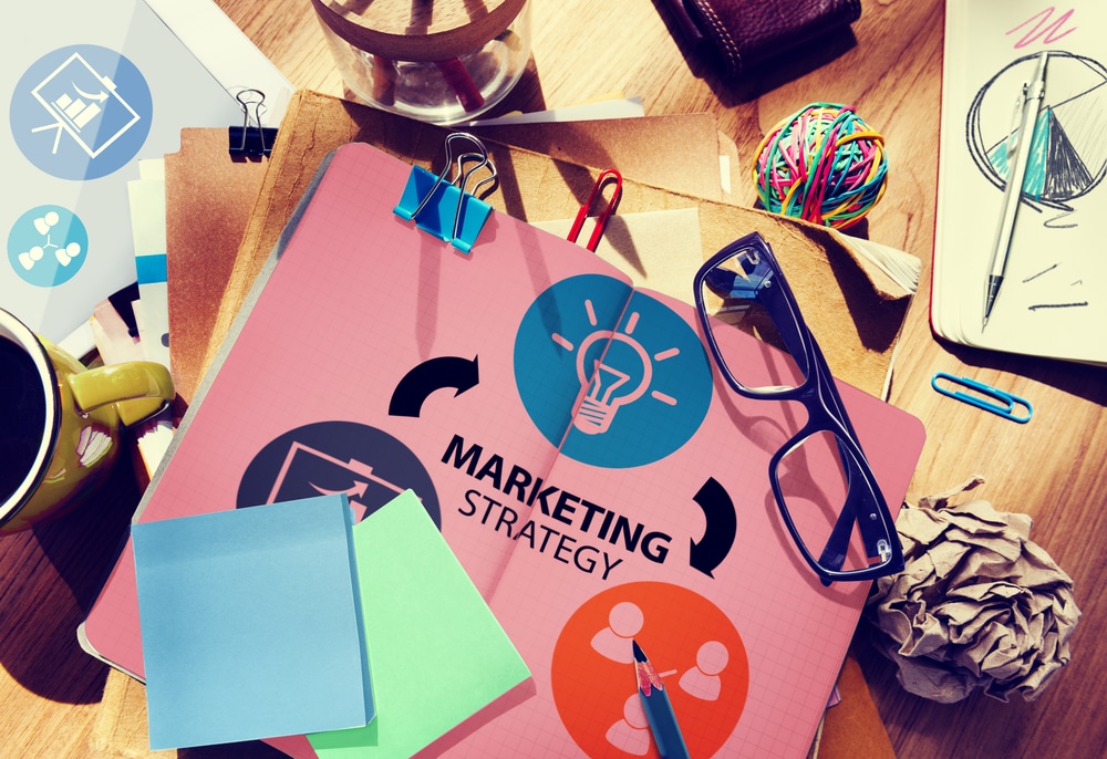 Zaśmiecone biurko z materiałami dotyczącymi strategii marketingowej, szklankami i filiżanką kawy – wskazuje na kreatywną sesję burzy mózgów.