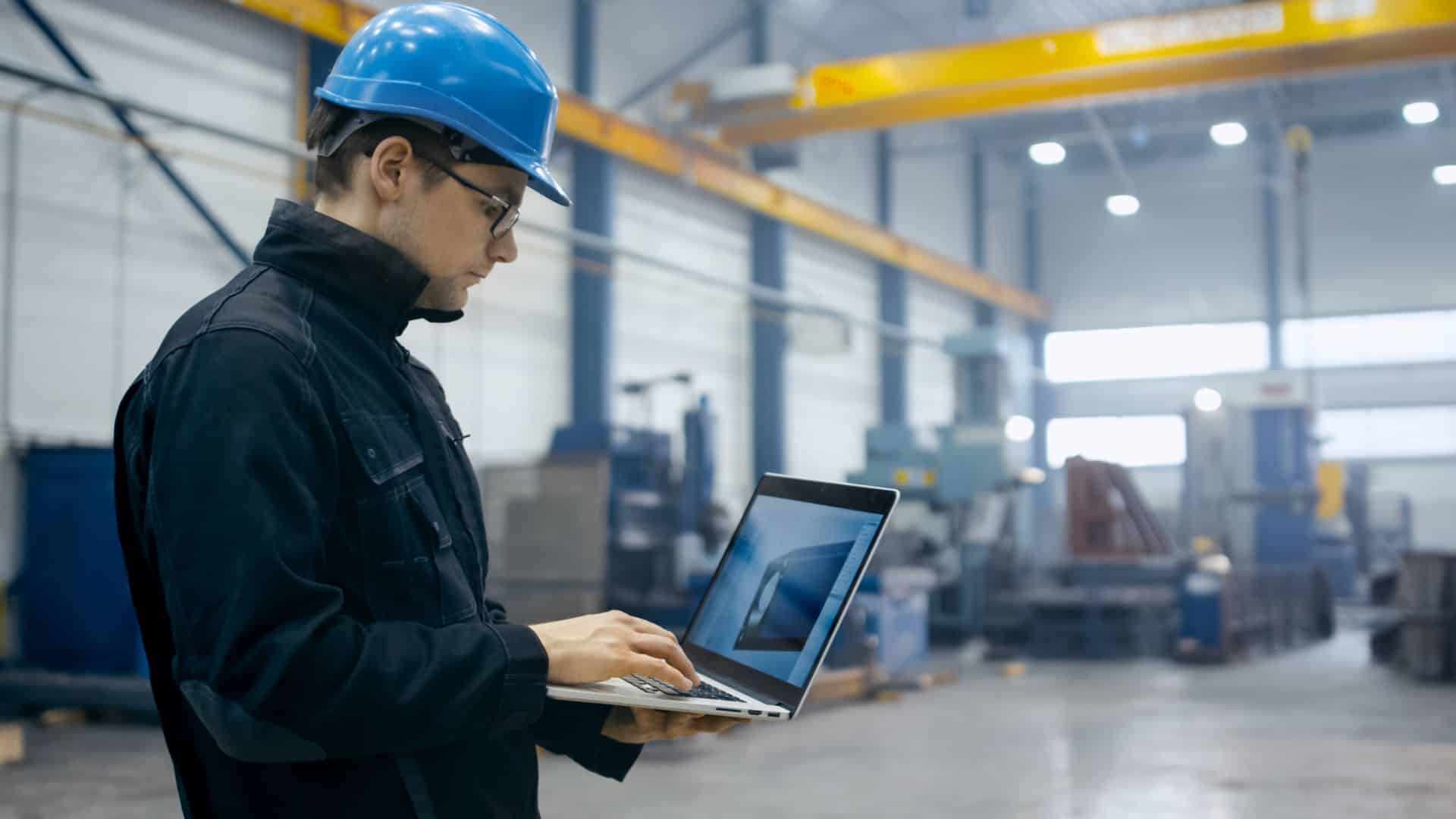 Inżynier w kasku i odzieży roboczej korzystający z laptopa w obiekcie przemysłowym.