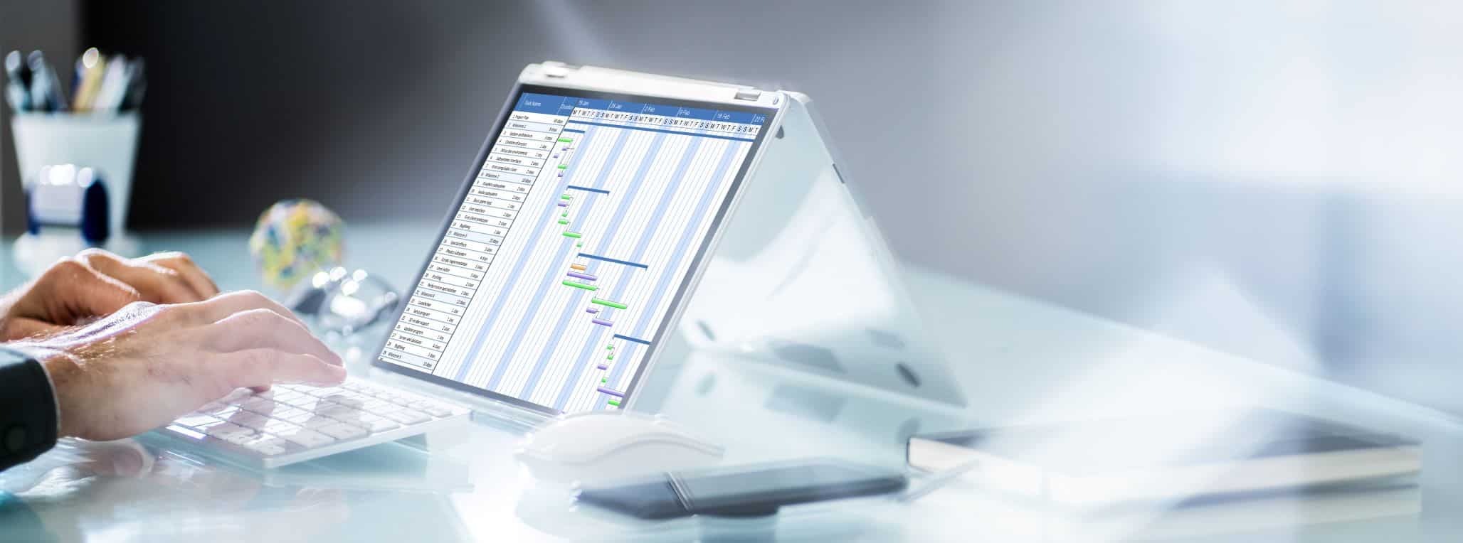 Osoba pracująca na laptopie wyświetlająca wykres Gantta z artykułami biurowymi i kulą ziemską w tle.