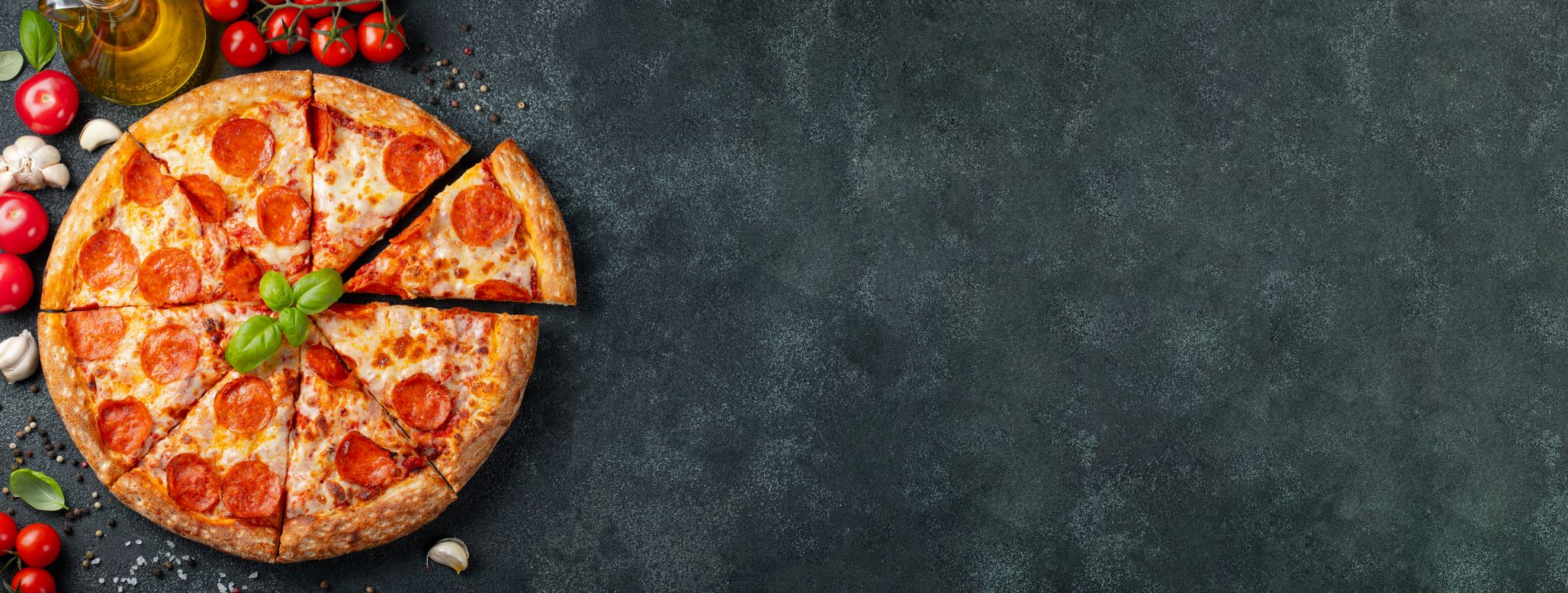 Pizza pepperoni, z której jeden kawałek został częściowo usunięty, jest umieszczana na ciemnej powierzchni o teksturze otoczonej takimi składnikami, jak pomidory, grzyby i oliwa z oliwek.