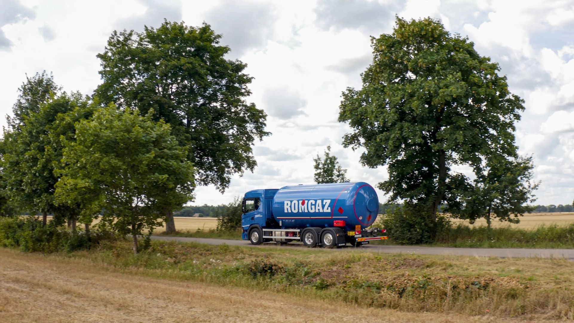 Niebieska cysterna Romgaz jedzie po wiejskiej drodze, otoczonej zielonymi drzewami i otwartymi polami uprawnymi pod zachmurzonym niebem.