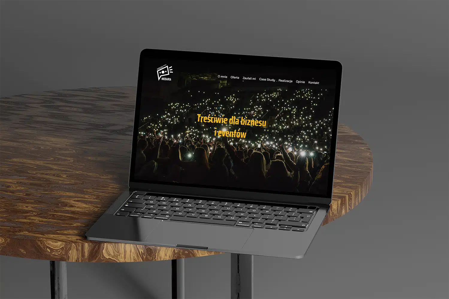 Laptop wyświetlający stronę internetową z wizerunkiem tłumu trzymającego podświetlane telefony, umieszczony na okrągłym drewnianym stole na szarym tle.
