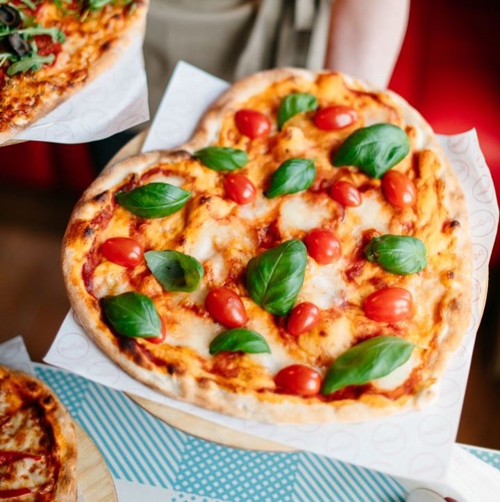 Pizza w kształcie serca, z pomidorkami koktajlowymi, liśćmi bazylii i roztopionym serem, położona na drewnianej desce pod niebiesko-białą matą.