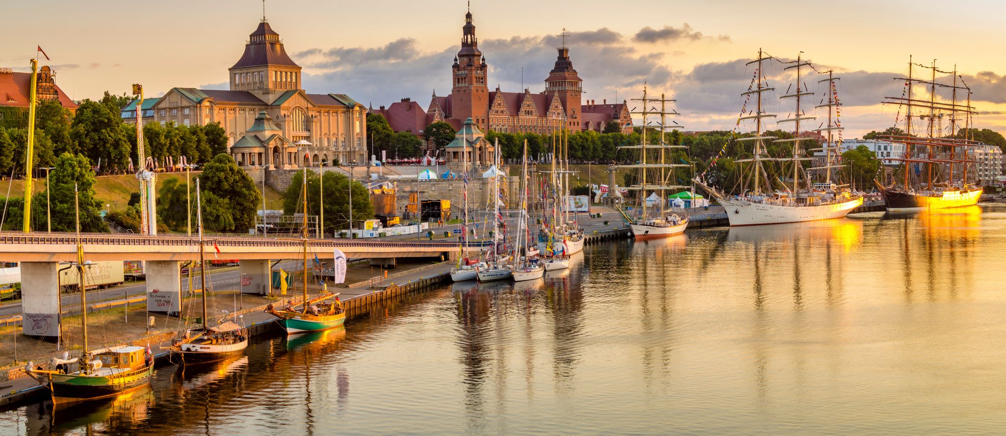 Widok na miasto o zachodzie słońca, z zabytkowymi budynkami, zacumowanymi żaglówkami i spokojnymi odbiciami wody.