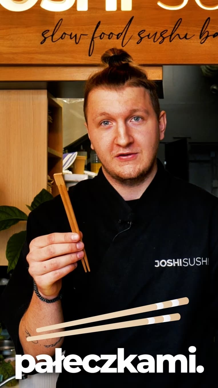 Osoba trzymająca pałeczki, ubrana w czarny uniform z napisem „JOSHI SUSHI”. Tło zawiera elementy kuchenne, a obraz ma tekst na dole z grafiką pałeczek.