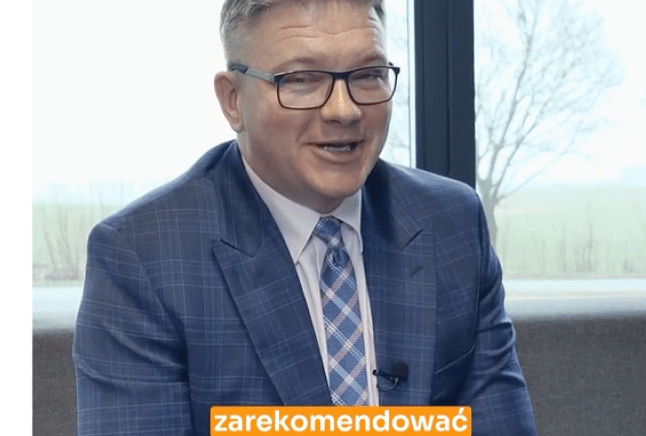 Mężczyzna w niebieskim kraciastym garniturze i wzorzystym krawacie siedzi w pomieszczeniu, uśmiecha się. Na dole nałożony tekst po polsku.