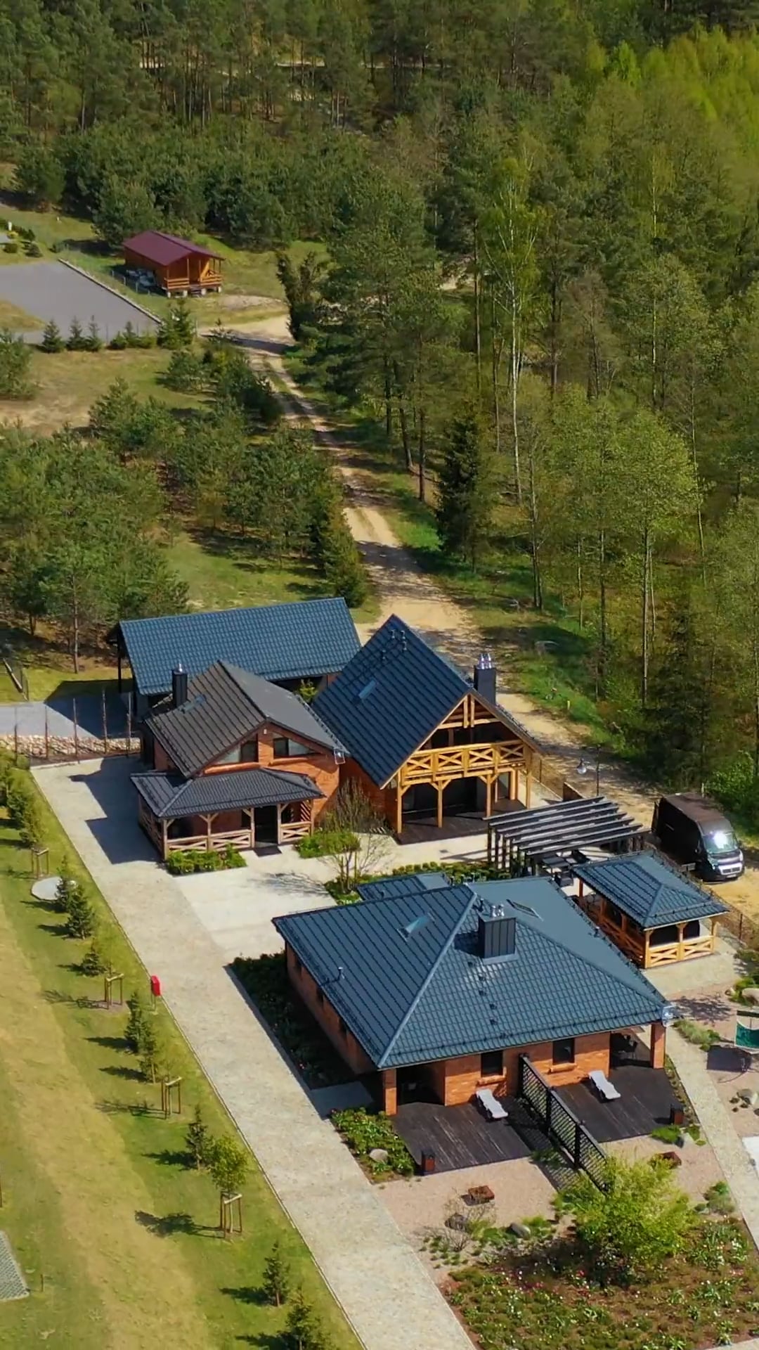 Widok z lotu ptaka na nowoczesną wiejską posiadłość z wieloma drewnianymi domami, niebieskimi dachami, podjazdem, zaparkowanym samochodem i otaczającymi drzewami.