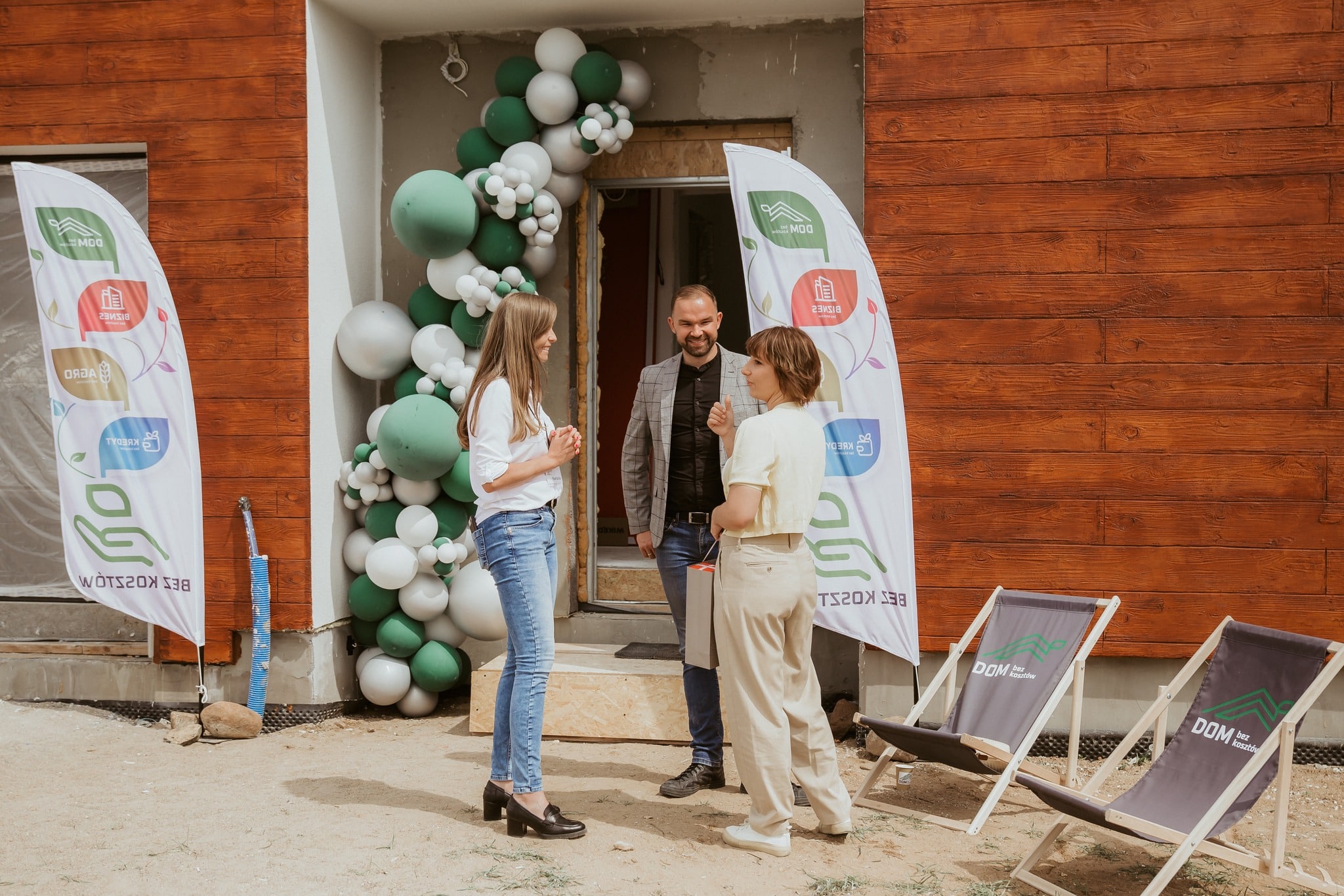 Trzy osoby stoją i rozmawiają przed udekorowanym balonami i banerami wejściem do budynku, w pobliżu dwóch złożonych leżaków na piaszczystym podłożu.