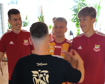 Czterech młodych mężczyzn stoi w środku; trzech nosi pasujące do siebie czerwone i żółte koszulki sportowe, podczas gdy jeden jest odwrócony w czarnej koszuli i gestykuluje rękami.