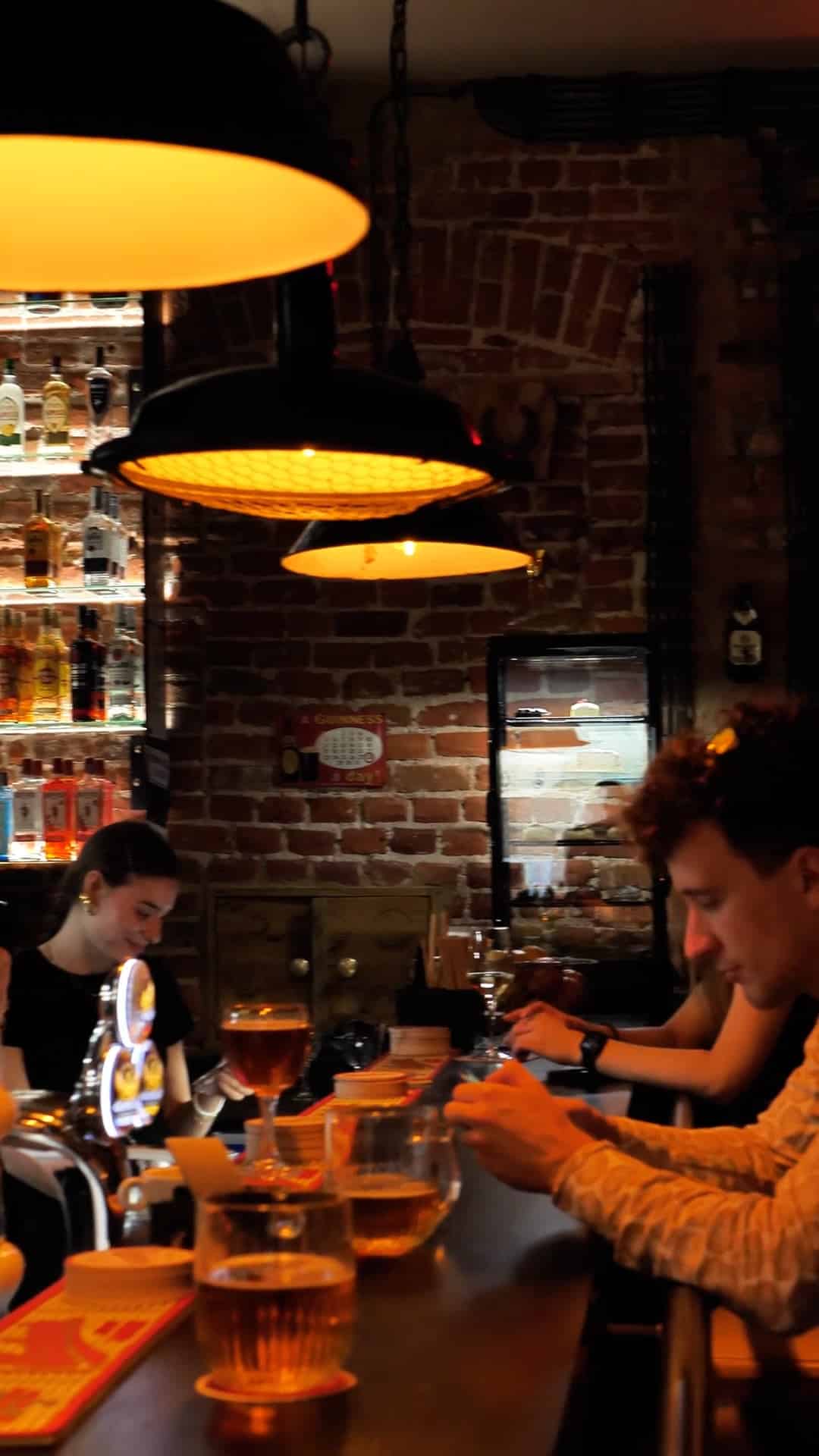 Ludzie siedzą przy barze z drinkami, oświetleni wiszącymi lampami, z barmanem pracującym za ladą i półkami z butelkami alkoholu w tle.