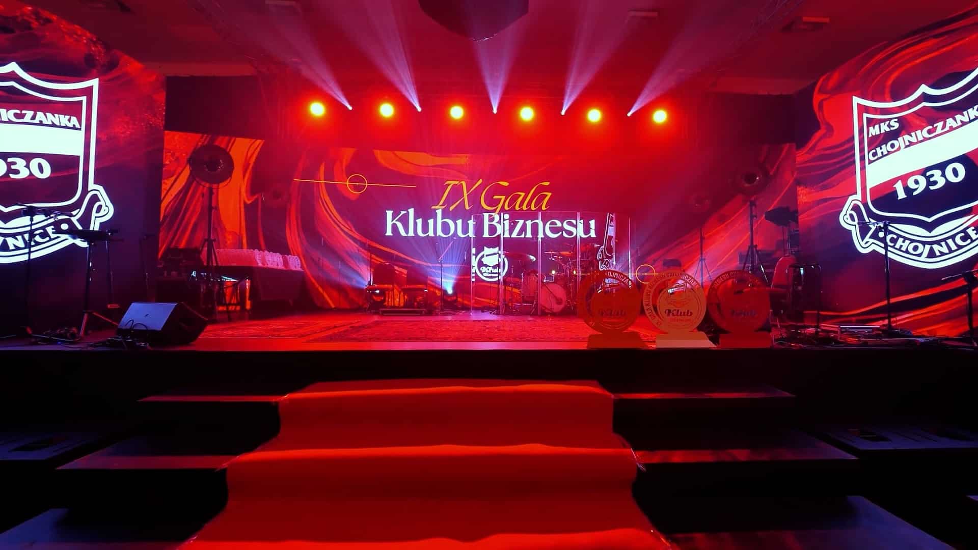 Scena z czerwonym oświetleniem i reflektorami jest przygotowana na "IX Galę Klubu Biznesu", z instrumentami muzycznymi i banerami eventowymi w tle.