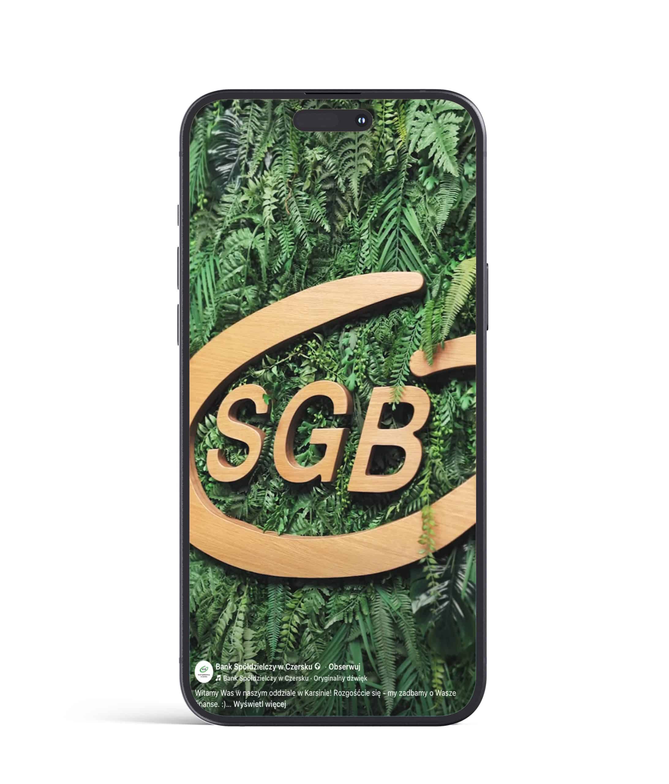 Smartfon wyświetlający zieloną ścianę roślin z dużym drewnianym logo SGB na ekranie.