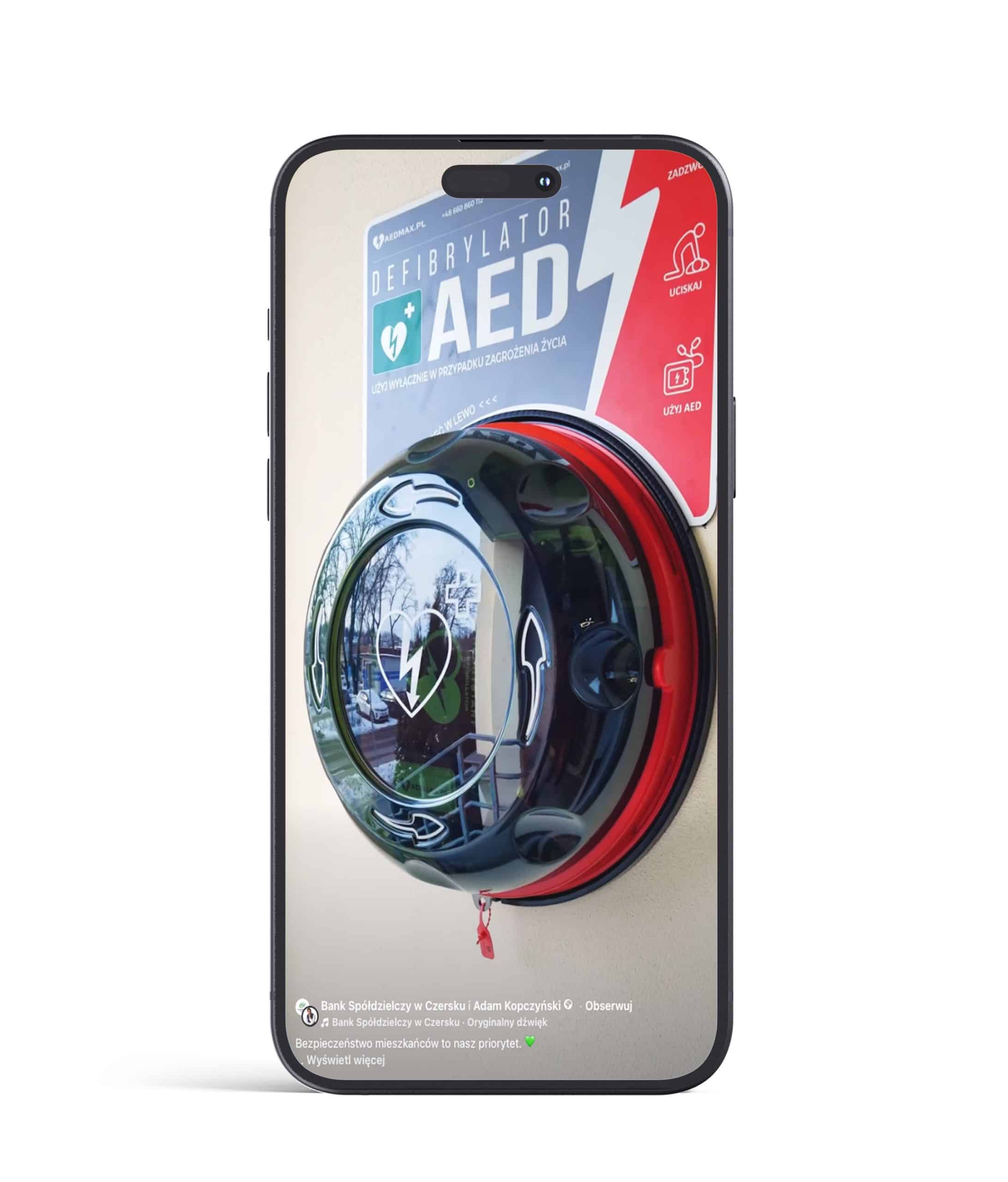Smartfon wyświetla zdjęcie zamontowanego na ścianie urządzenia AED (automatycznego defibrylatora zewnętrznego) w okrągłej obudowie z napisem "Defibrylator AED".